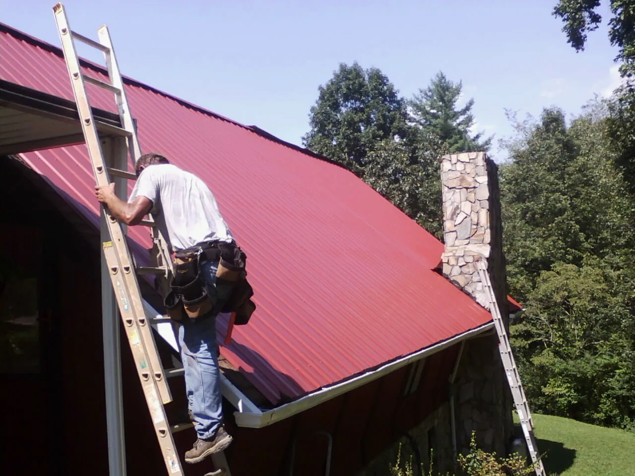 Roofer on a ladder beside a red metal roof and stone chimney — Soffit & Fascia Repair by Roofing Brothers in Cuyahoga Falls